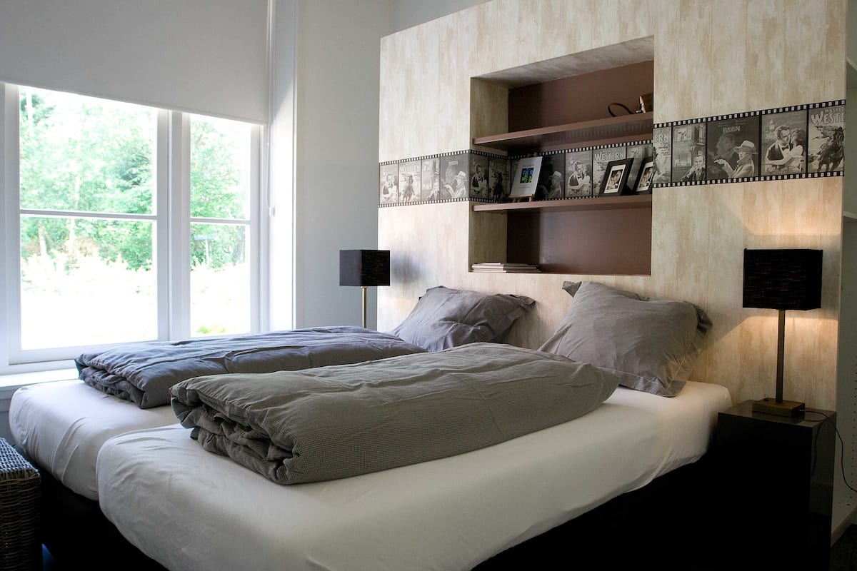 A bedroom with two beds, a large window, and a wall-mounted shelf with framed pictures.