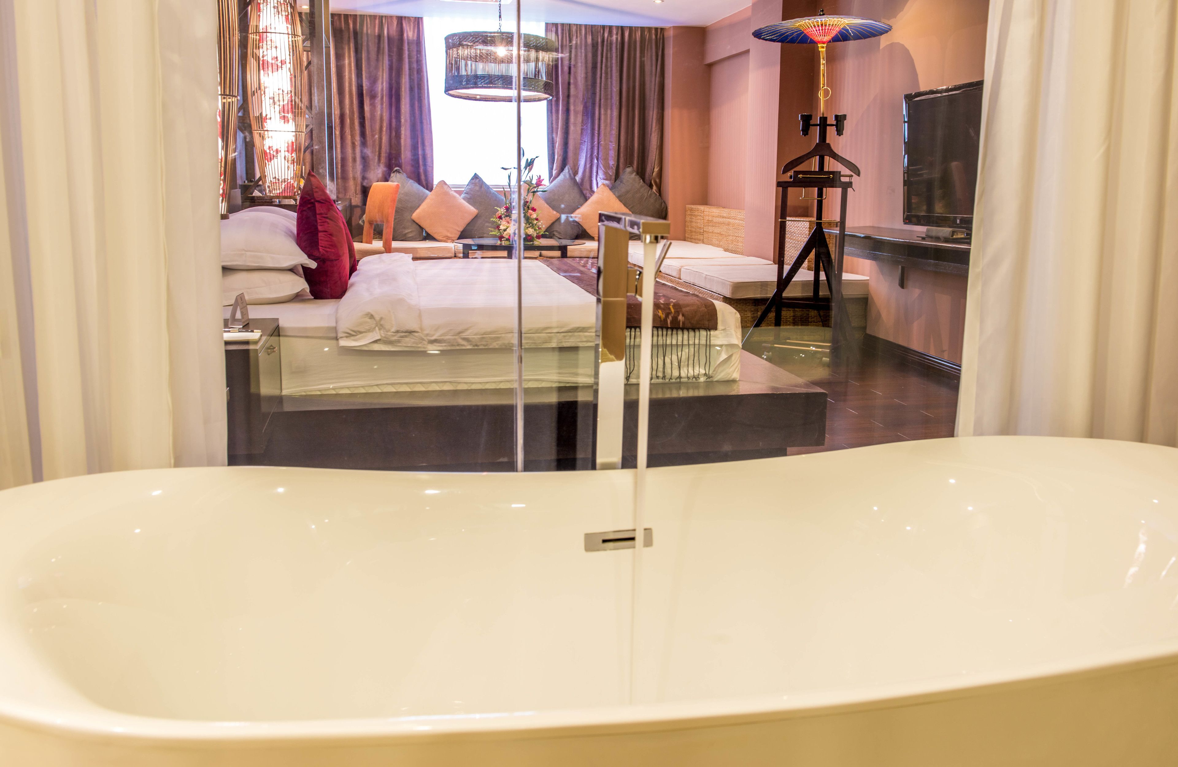 Una habitación de hotel moderna con una bañera grande, una cama con muchas almohadas, un televisor y una lámpara de pie.