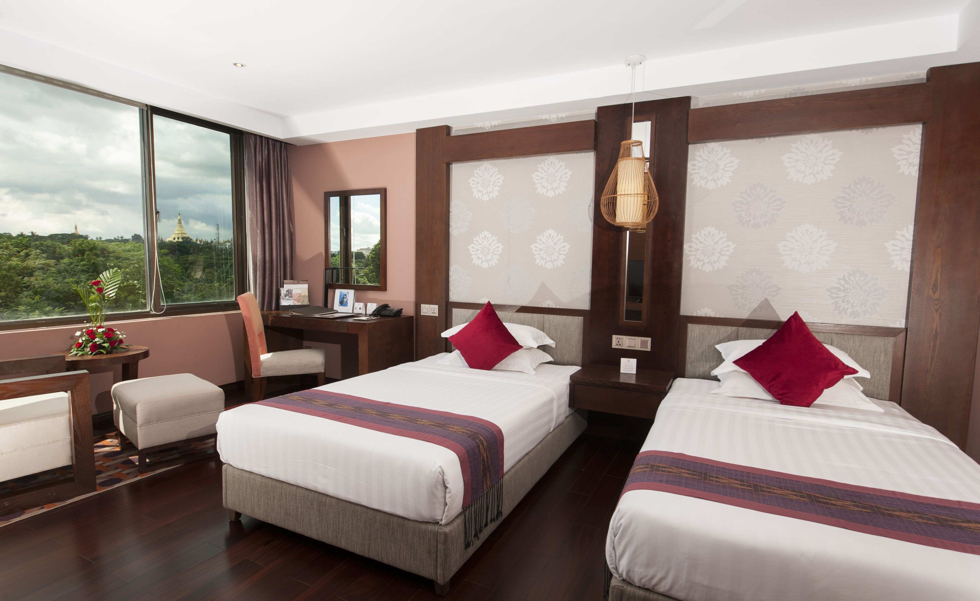 A hotel room with two beds, a desk, a chair, and a large window offering a view of greenery.