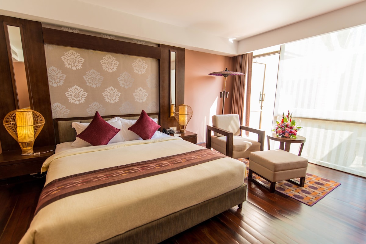 A hotel room with a large bed, a chair, a small table with flowers, and a view of the outdoors.