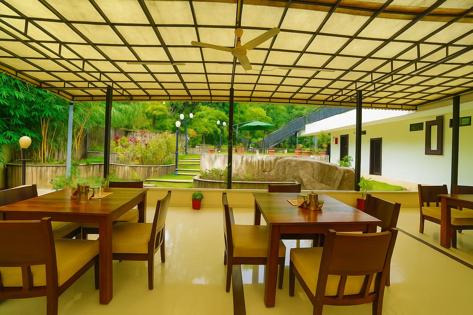 A covered outdoor dining area with wooden tables and chairs, a ceiling fan, and a view of a garden with a stone wall and steps.