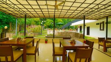 A covered outdoor dining area with wooden tables and chairs, a ceiling fan, and a view of a garden with a stone wall and steps.