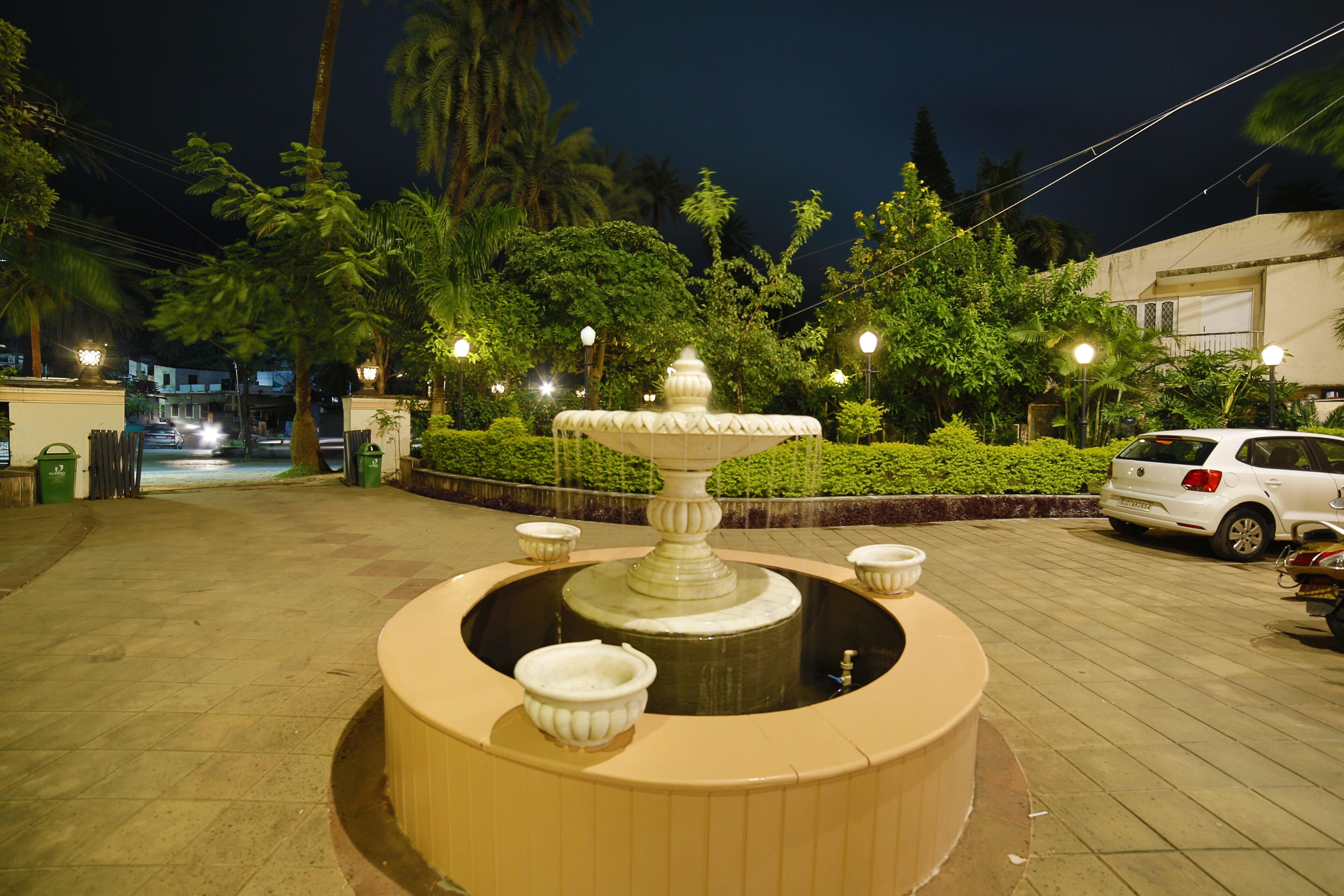 A circular fountain with a central pedestal and surrounding bowls, surrounded by well-maintained greenery and street lamps.