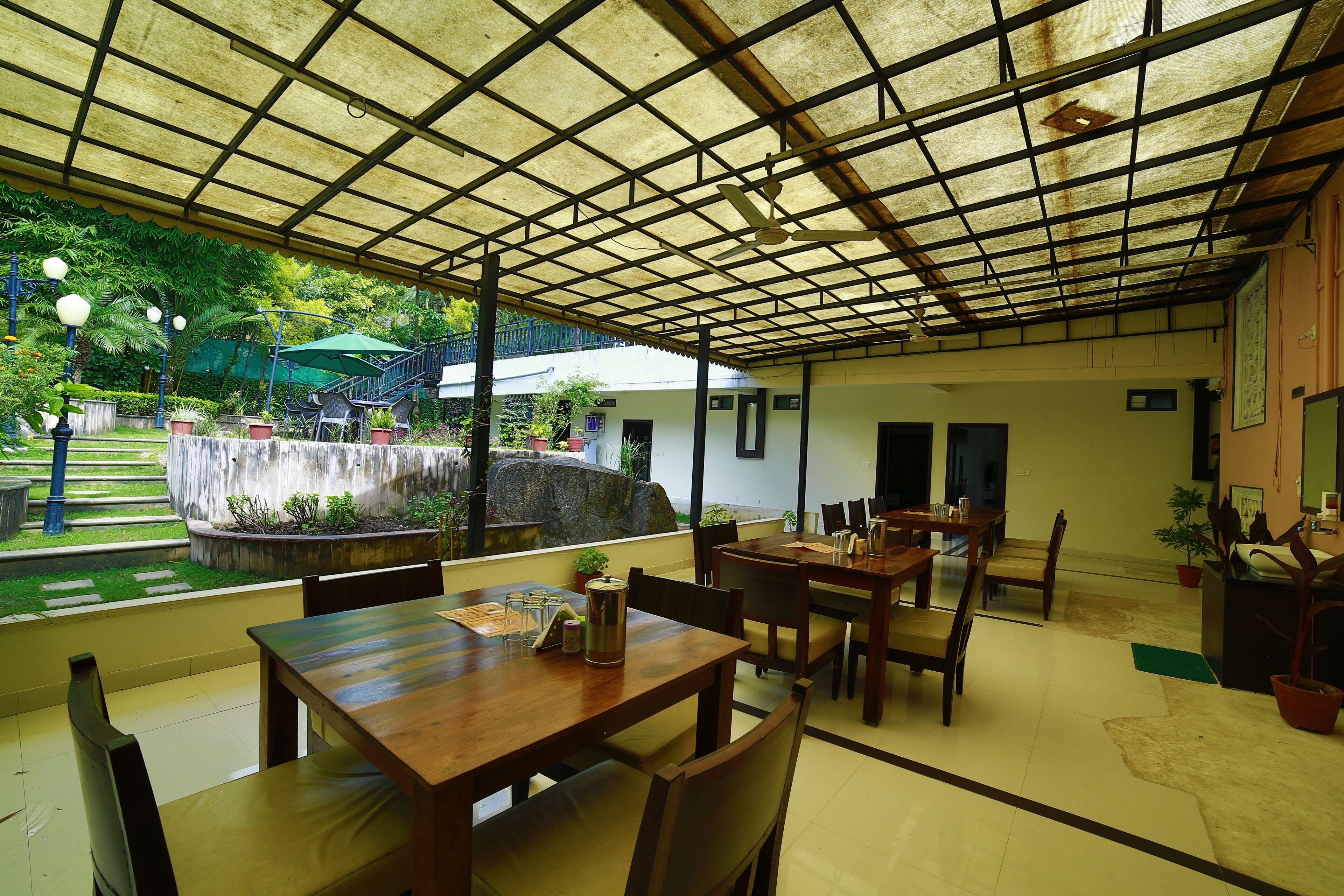 Un espace de restauration intérieur avec des tables et des chaises en bois, un ventilateur de plafond et une vue sur un jardin agrémenté d’un élément aquatique et de plantes.