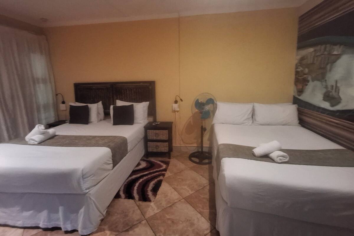A hotel room with two single beds, a nightstand, a wall-mounted fan, and a painting.