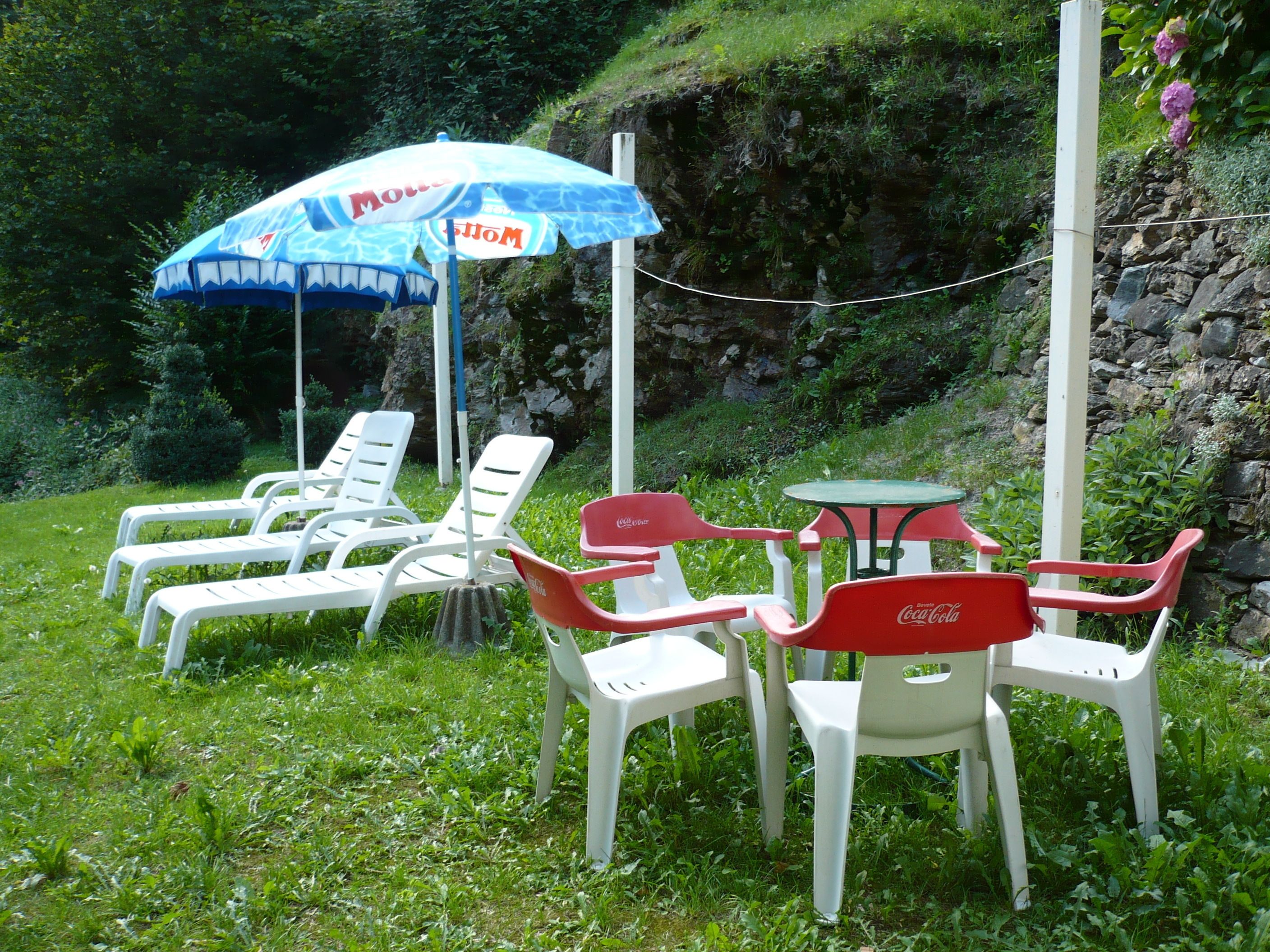 Mobiliario exterior con sillas blancas y rojas y una mesa, rodeado de follaje y un muro de piedra.