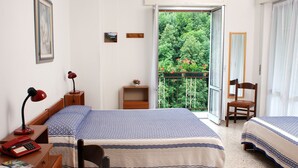 A bedroom with two beds, a desk with a typewriter, a chair, a small table with a lamp, and a balcony with a view of greenery. - Albergo Regina (Bognanco)