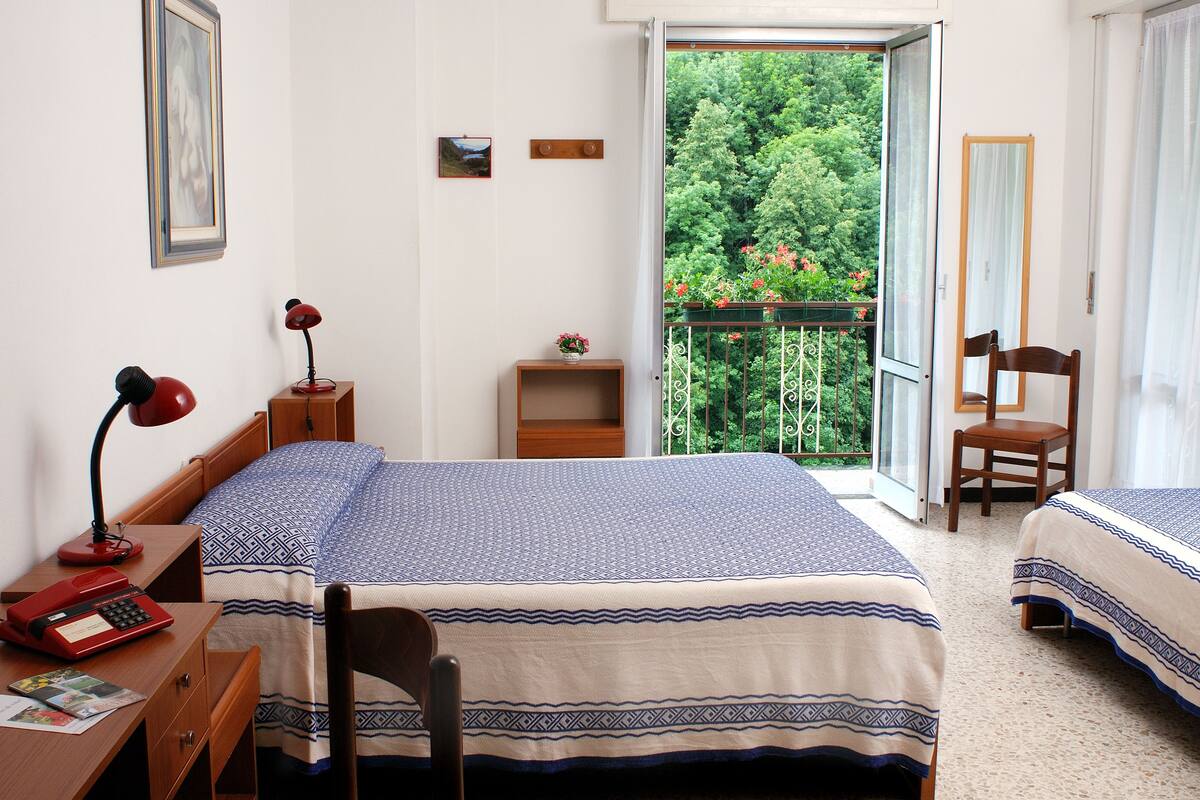 A bedroom with two beds, a desk with a typewriter, a chair, a small table with a lamp, and a balcony with a view of greenery.