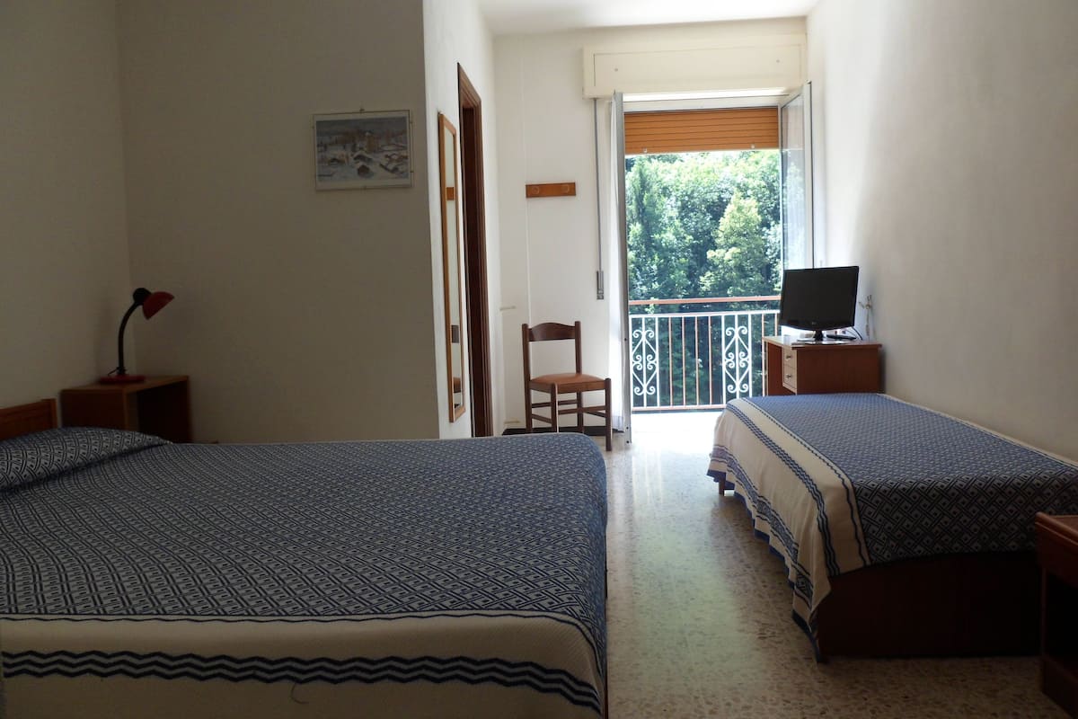 A room with two beds, a chair, a desk with a computer, and a balcony with a railing.