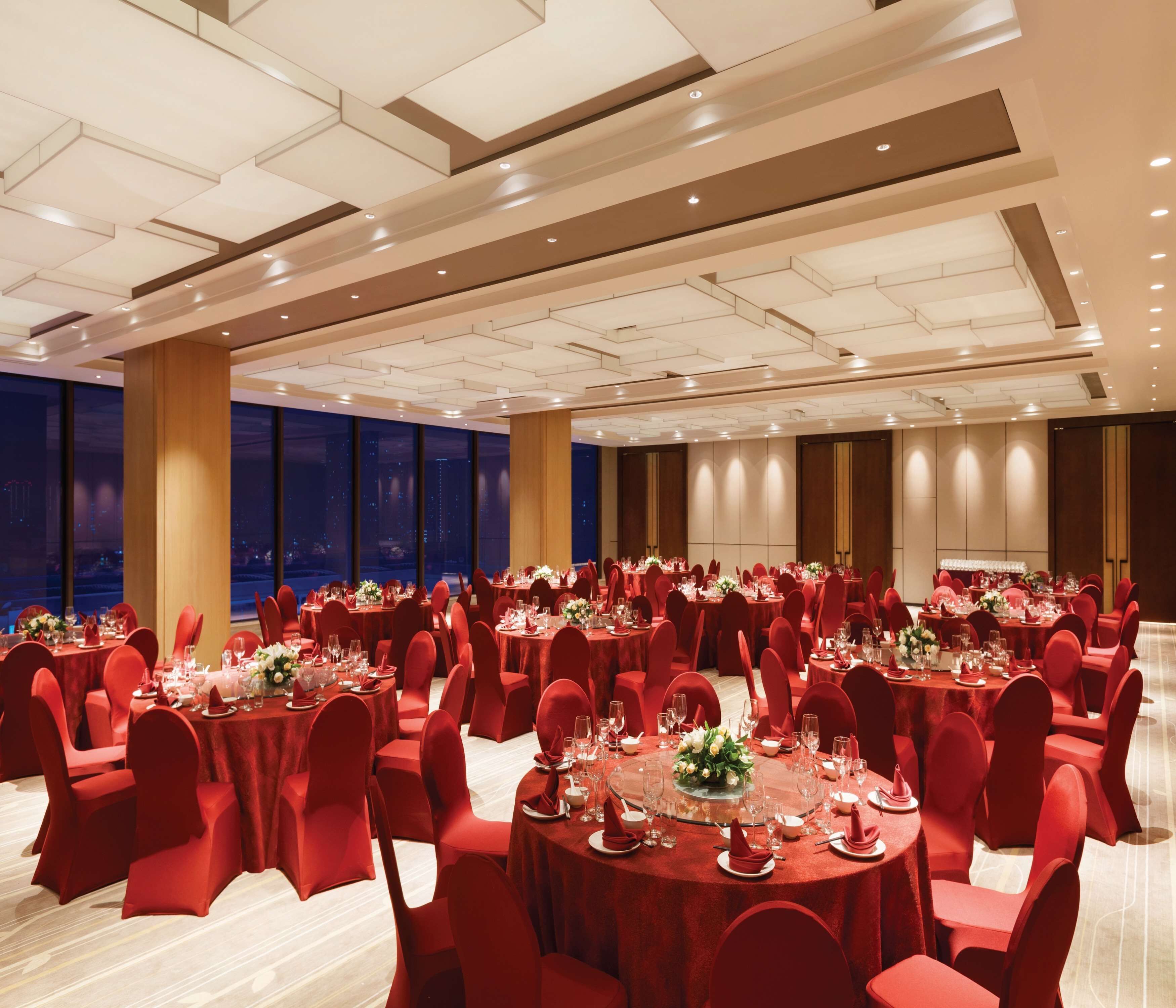 Une salle de banquet avec des tables rondes, des chaises rouges et une vue sur la ville à travers de grandes fenêtres.