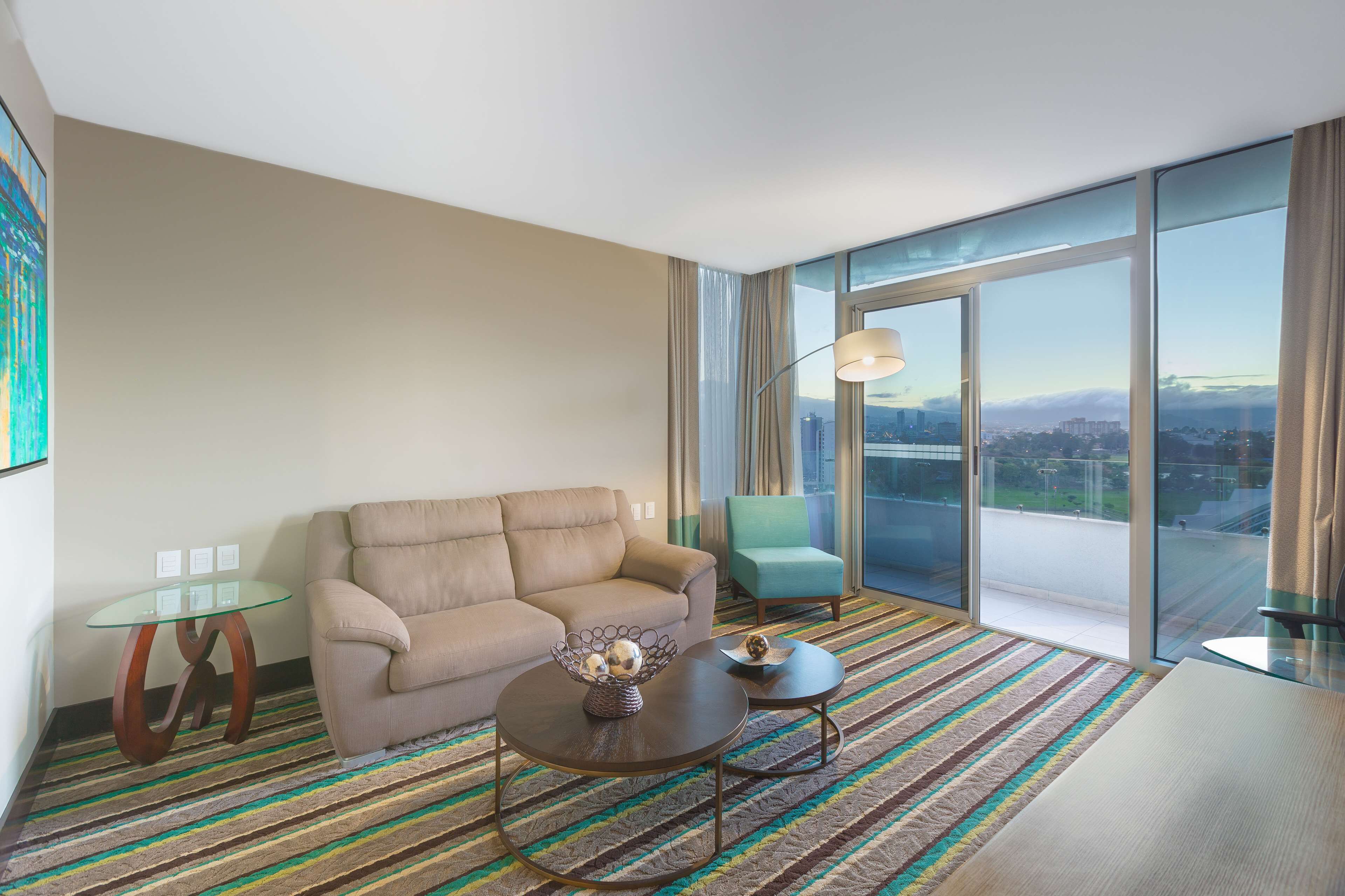A modern hotel room with a sofa, two coffee tables, a glass table, a green armchair, and a large window offering a city view.