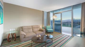 A modern hotel room with a sofa, two coffee tables, a glass table, a green armchair, and a large window offering a city view.
