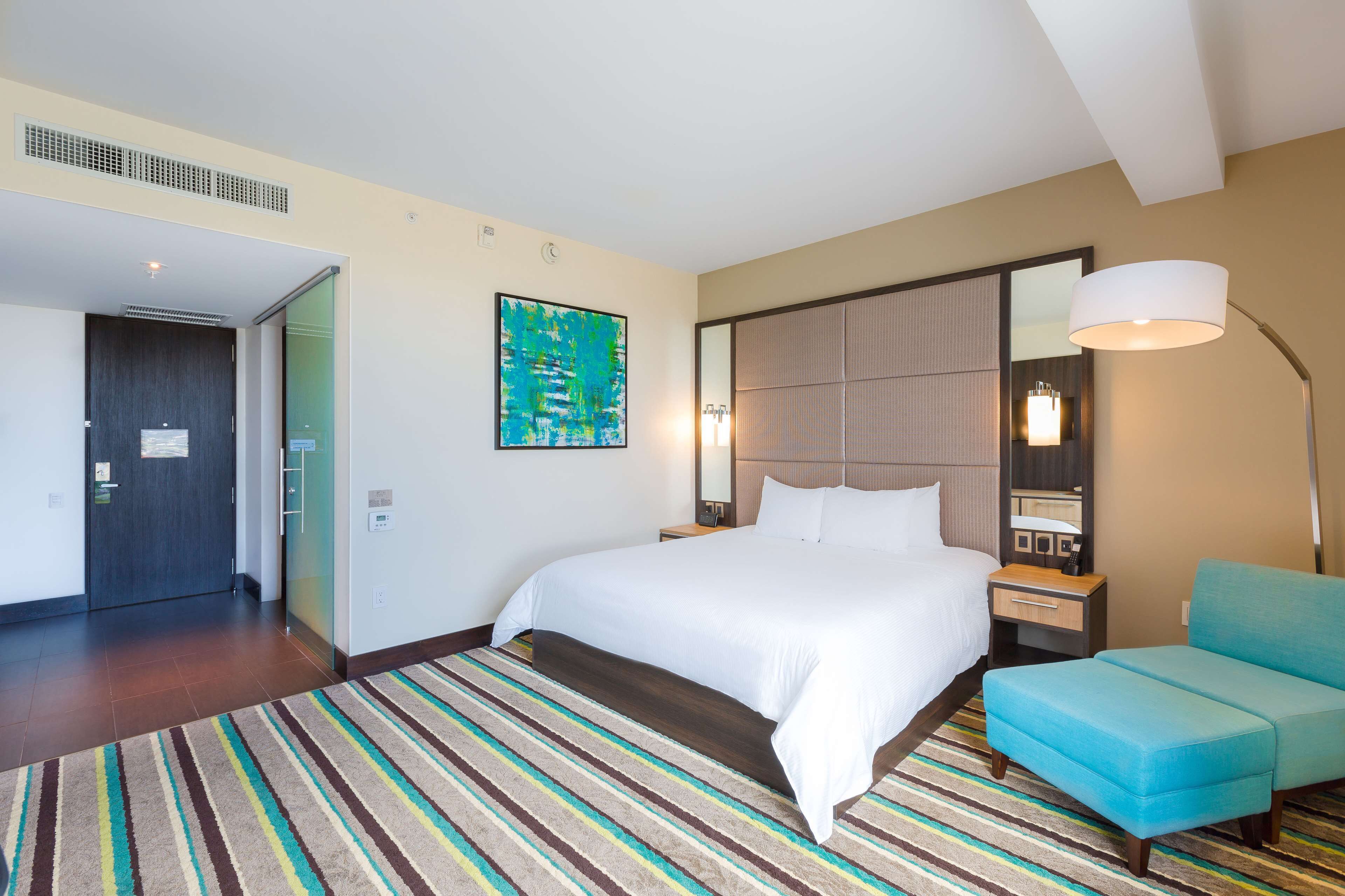 A hotel room with a large bed, a nightstand, a desk, a chair, a large mirror, and a colorful striped rug.