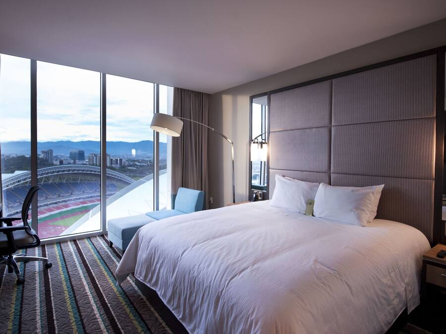 Quarto de hotel com uma cama grande, uma escrivaninha, uma cadeira e vista para um estádio através de amplas janelas.