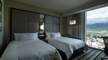 A hotel room with two beds, a large window with a city view, and a view of mountains.