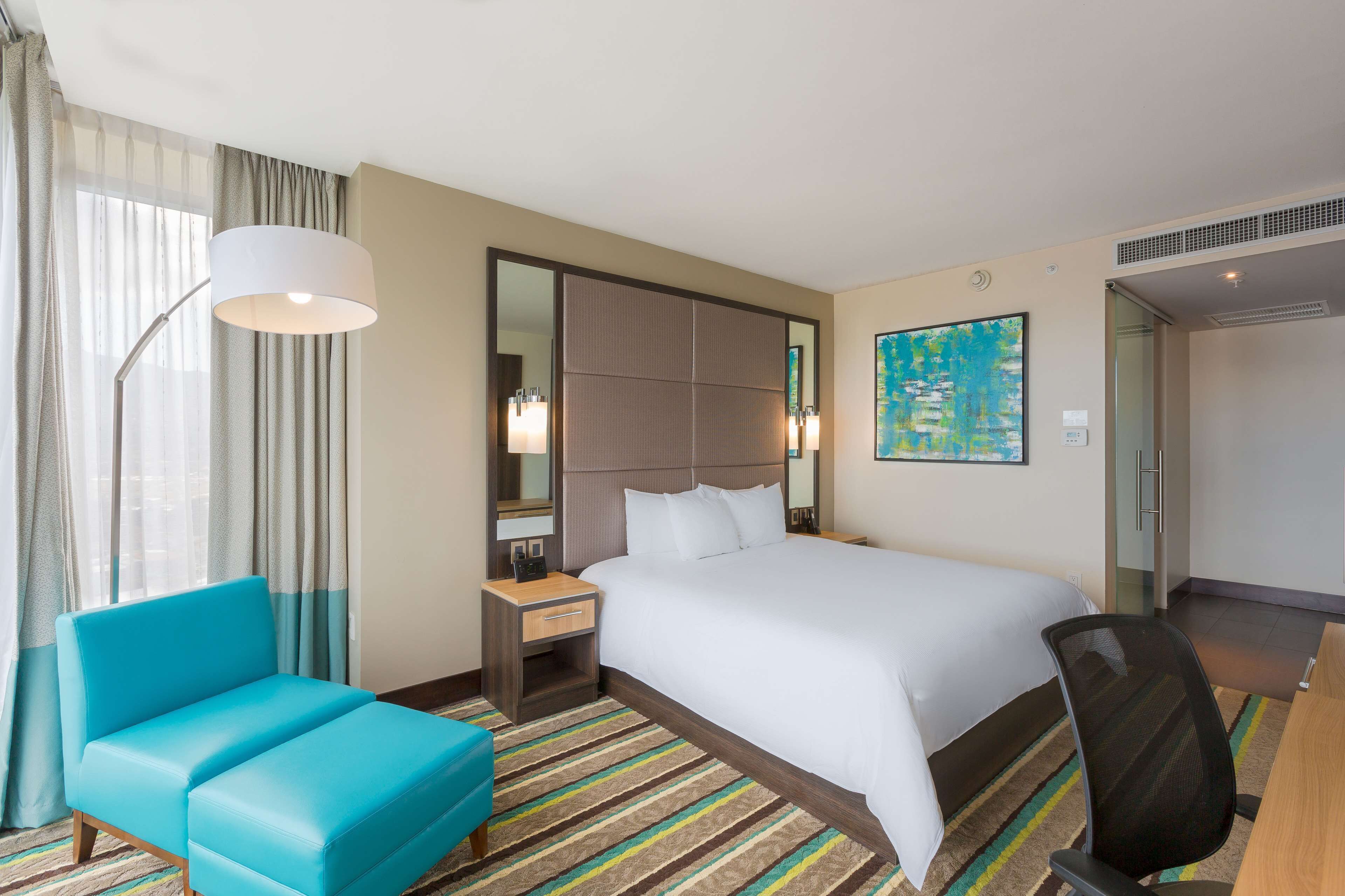 A hotel room with a large bed, a desk, a chair, a lamp, and a painting on the wall.