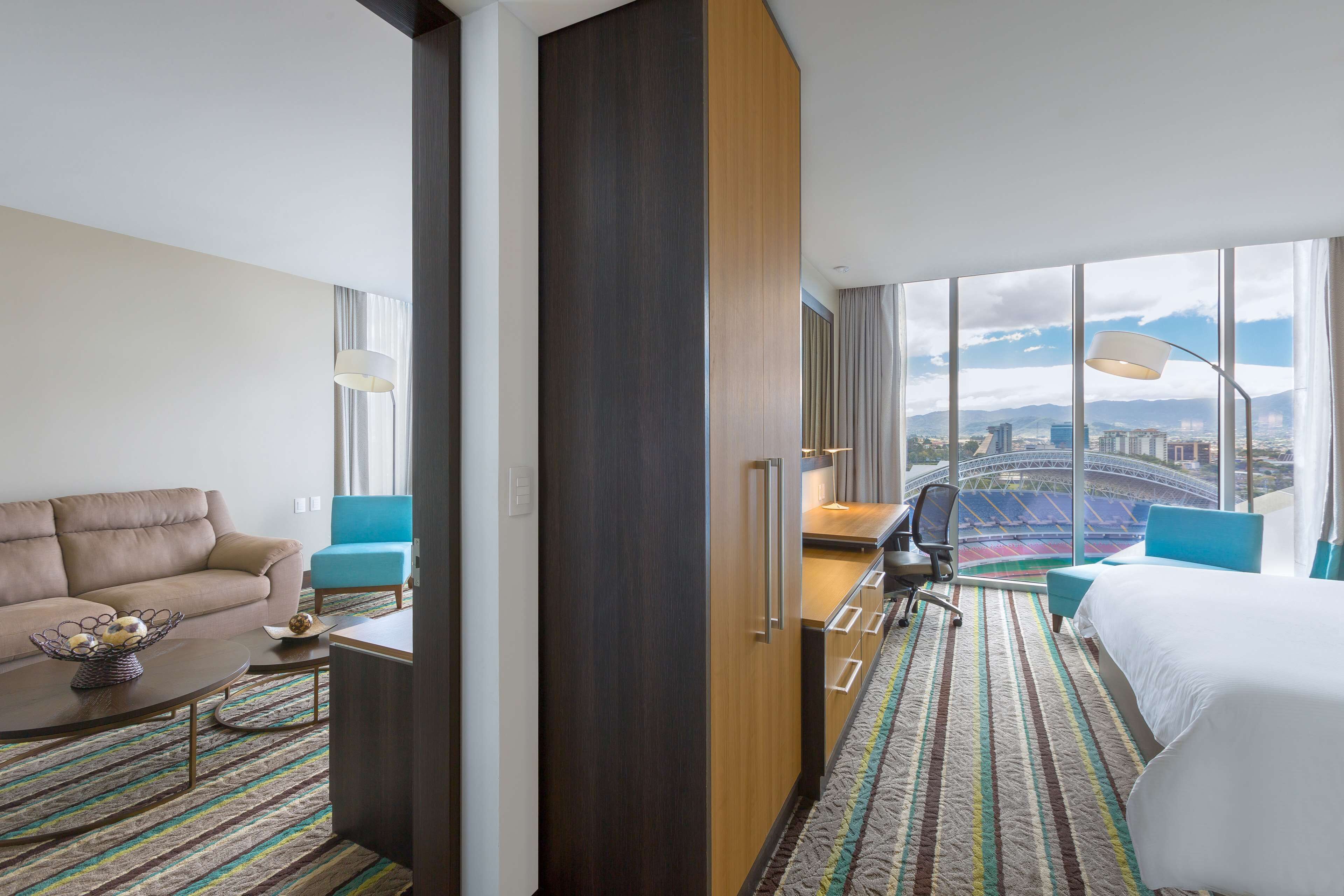A hotel room with a large bed, a sofa, a desk, and a view of a cityscape.