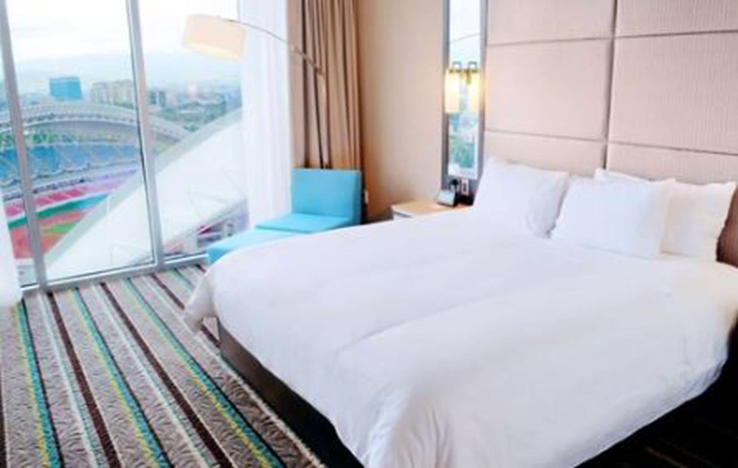 Een hotelkamer met een groot bed en een uitzicht op een stadion door middel van ramen van vloer tot plafond, plus een blauwe stoel.