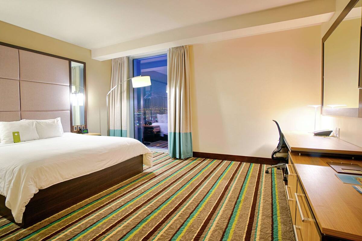 Hotelkamer met groot bed, bureau met stoel, raam met gordijnen en uitzicht op de stad 's nachts.