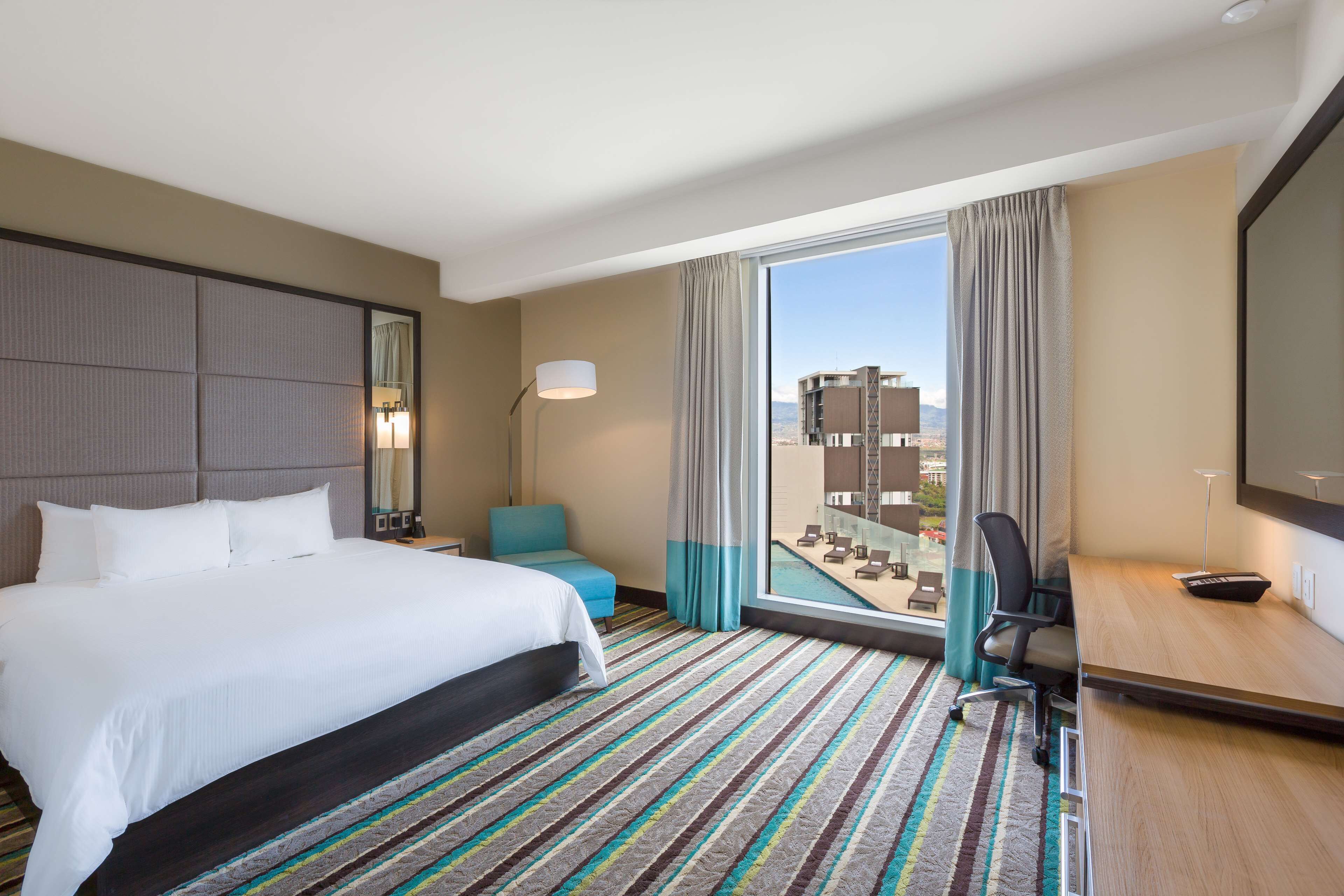 Habitación de hotel con una cama grande, un escritorio, una silla, vistas a la ciudad y un balcón con piscina.