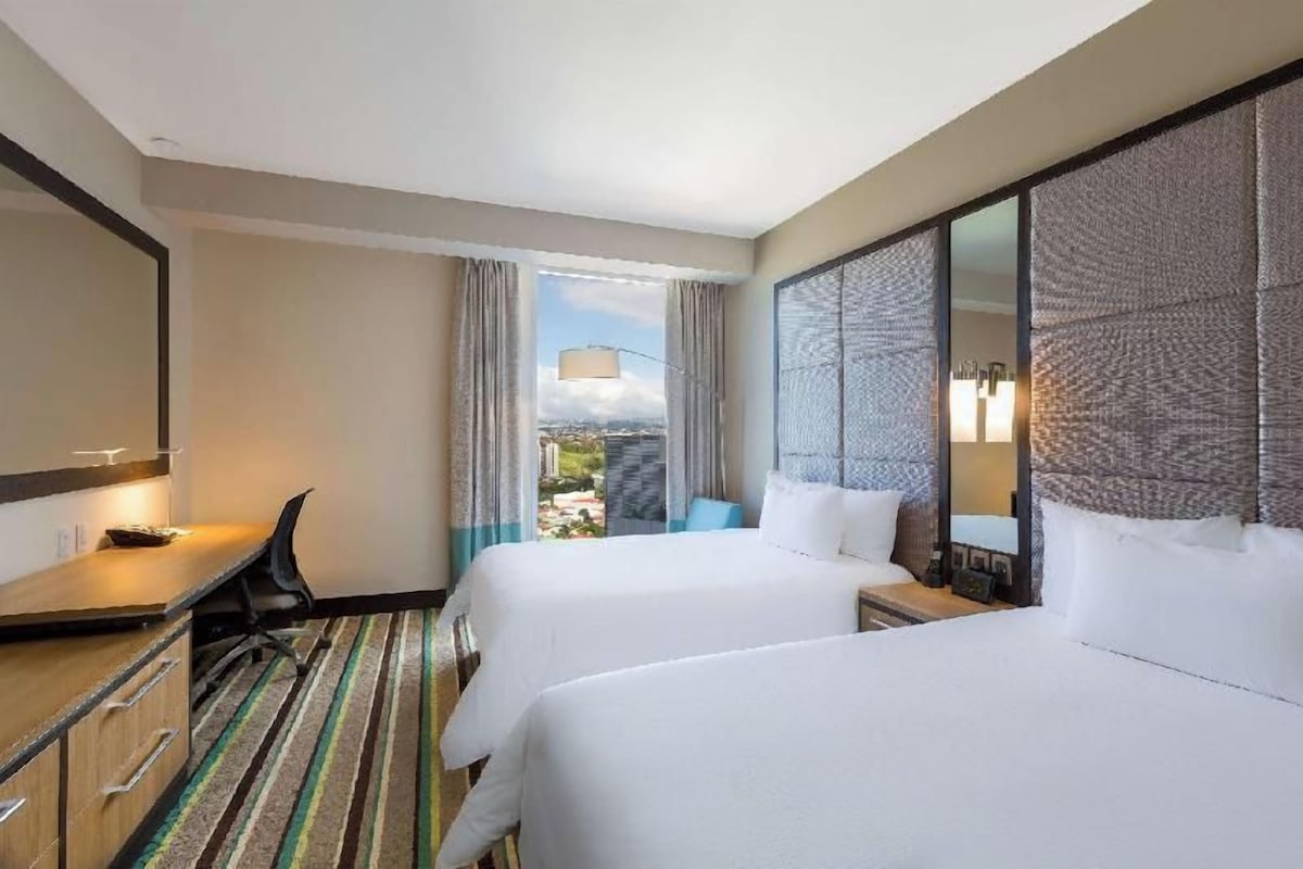 Une chambre d’hôtel avec deux lits, un bureau, une chaise, un grand miroir et une vue sur l’extérieur.