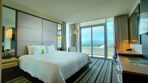 Habitación de hotel con una cama grande, un escritorio, una silla y vistas a la ciudad a través de las ventanas.