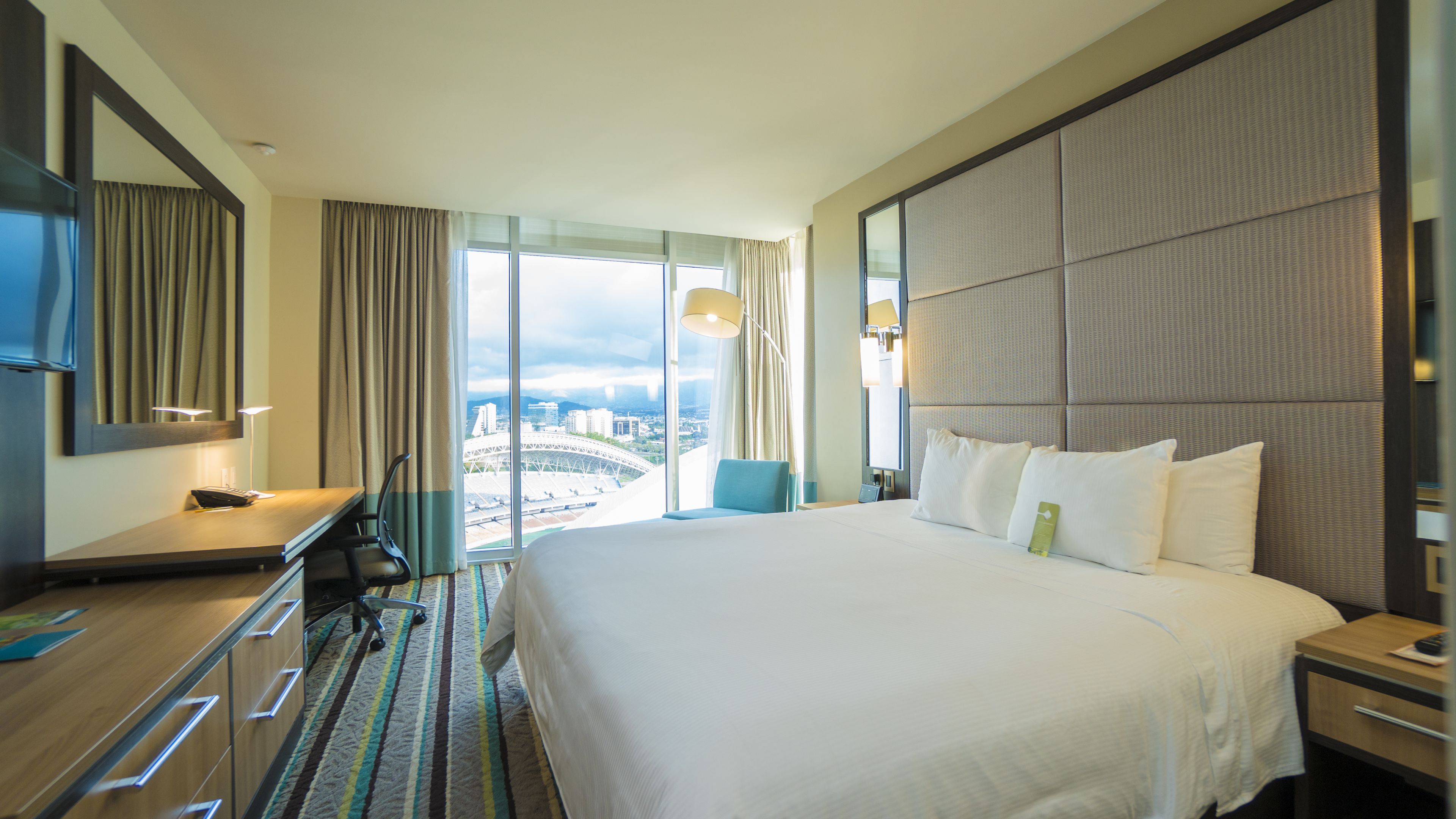 A hotel room with a large bed, a desk with a chair, a TV, and a view of the cityscape through large windows.