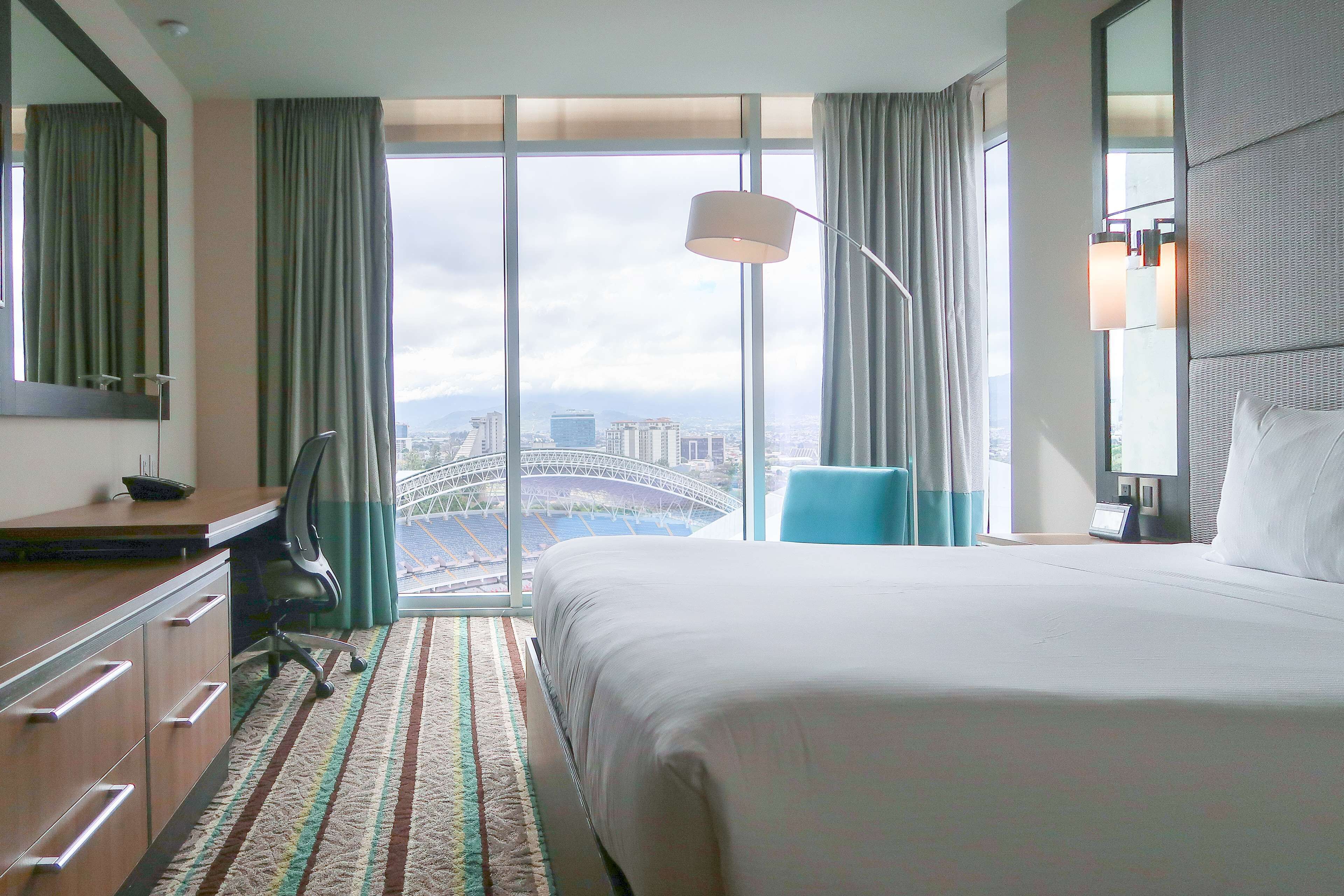 A hotel room with a large bed, a desk with drawers, a chair, a lamp, and a view of a cityscape through large windows.