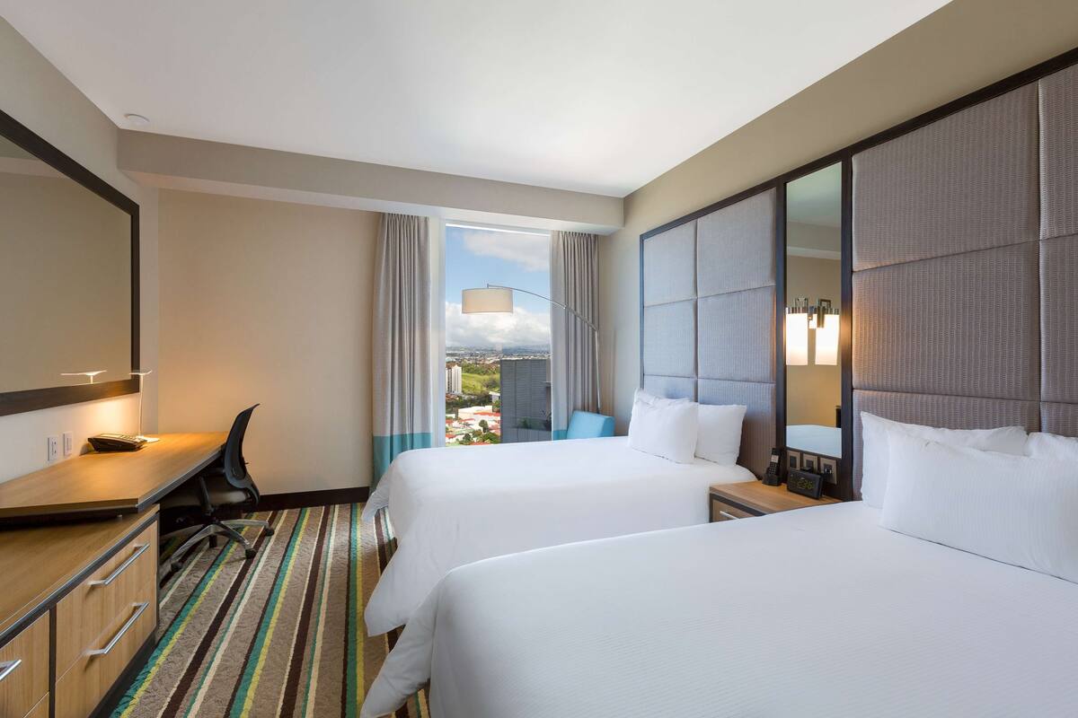 Een hotelkamer met twee bedden, een bureau, een stoel en een groot raam met uitzicht op de stad.