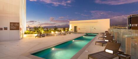 Zona de piscina con sillones de descanso y vistas a la ciudad al atardecer.