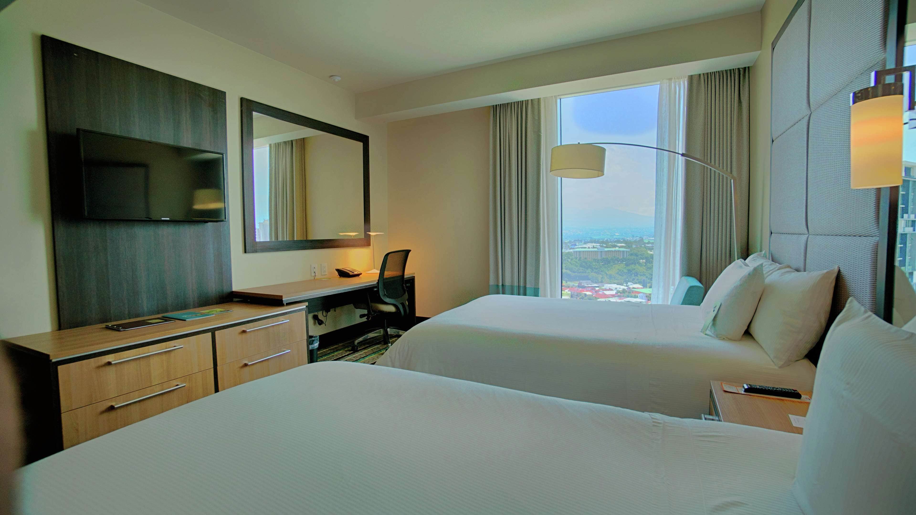 A hotel room with two beds, a desk, a chair, a TV, and a large window with a view.