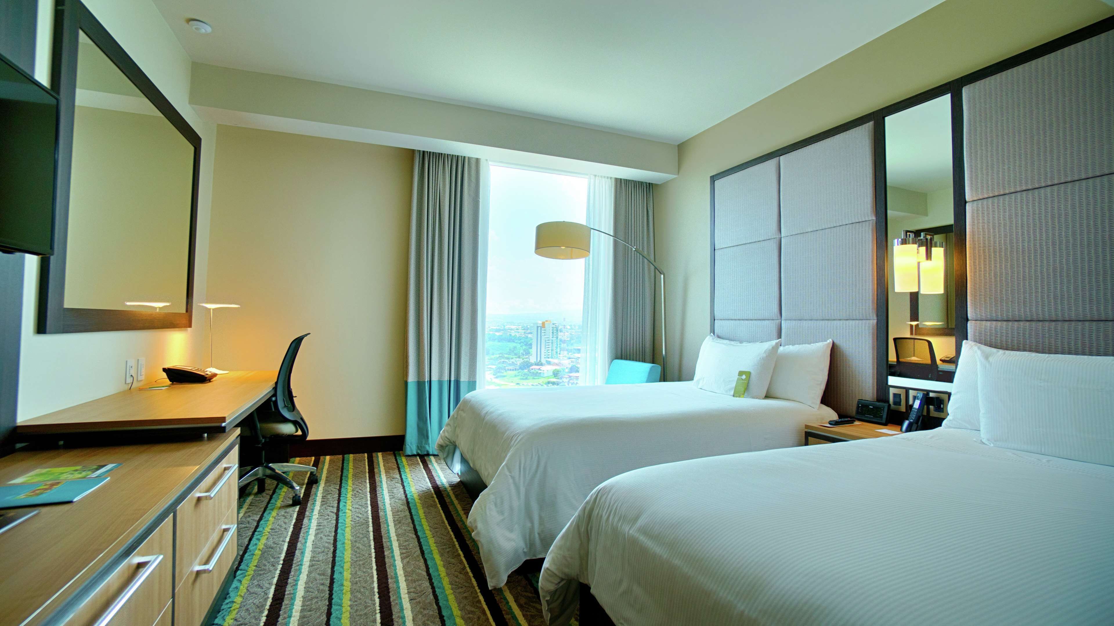 A hotel room with two beds, a desk, a chair, a large mirror, and a view of the city through the window.