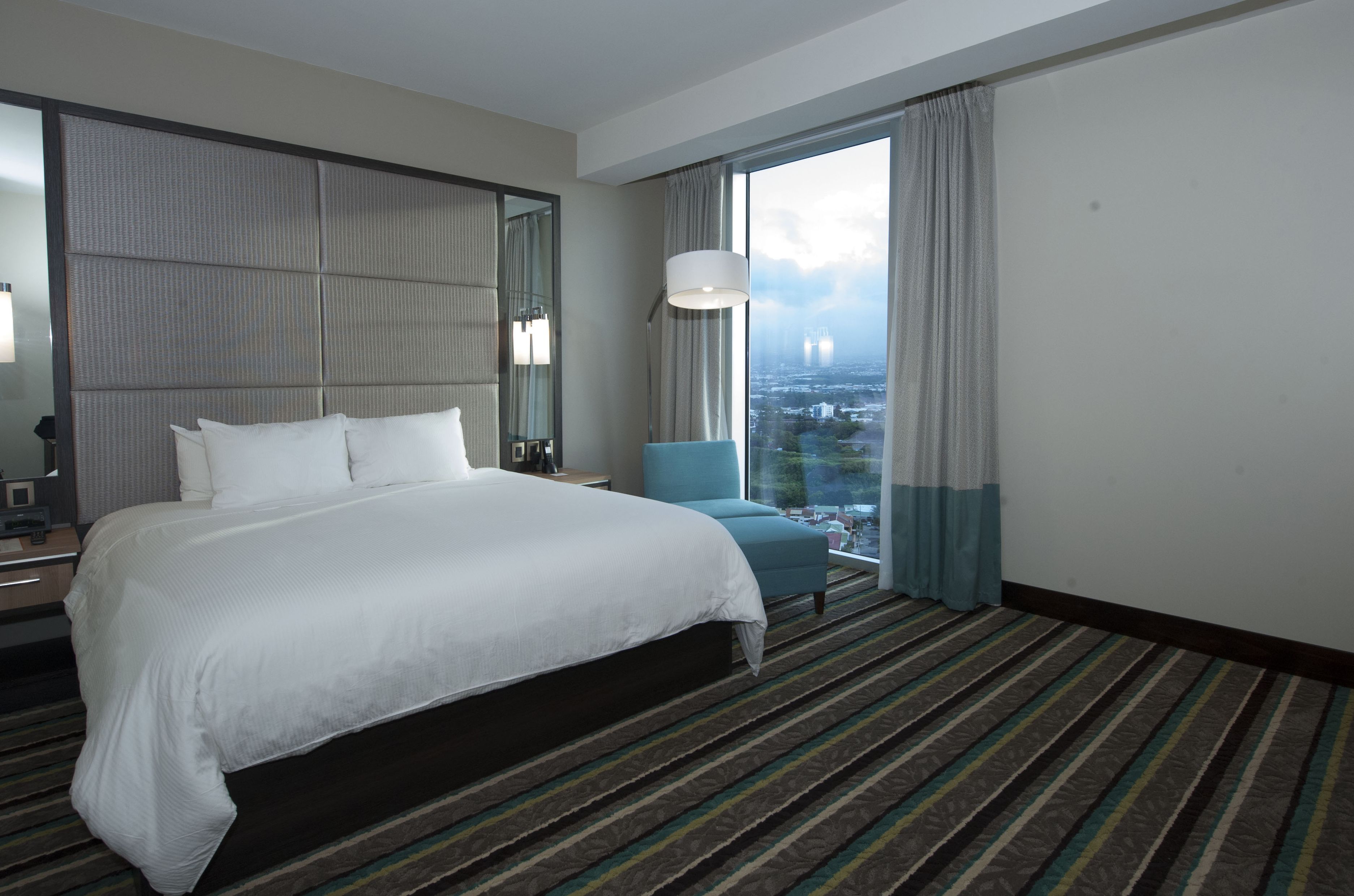 Ein Hotelzimmer mit einem großen Bett, einem Sessel, Blick auf die Stadt und einem großen Fenster.