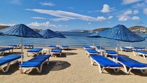 A beach with rows of sun loungers and blue umbrellas, a clear sky, and calm sea. - Nagi Beach Hotel (Bodrum)