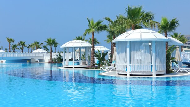 A swimming pool with clear blue water, surrounded by palm trees and white cabanas. - Sueno Hotels Deluxe Belek - All Inclusive (Serik)
