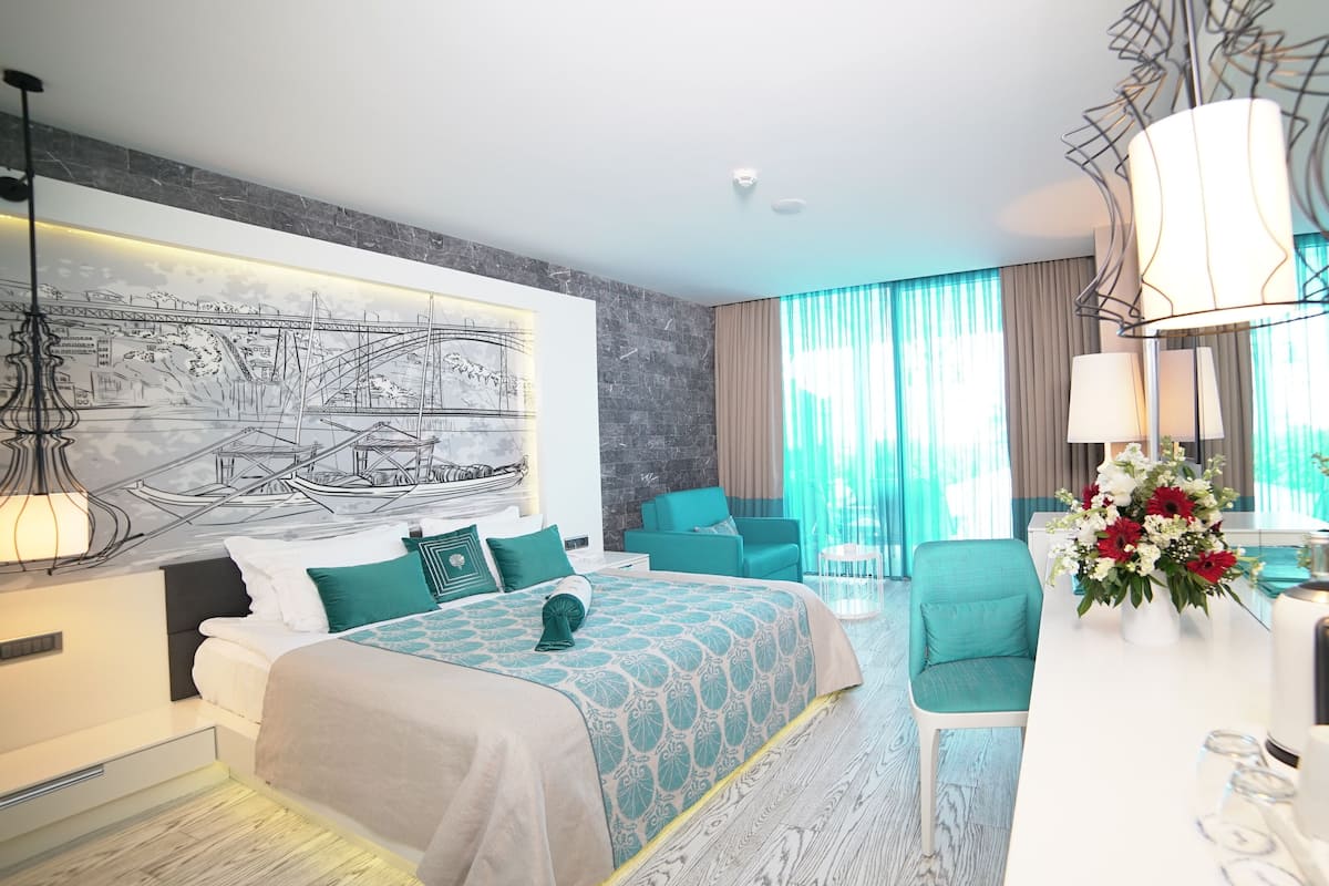Ein modernes Hotelzimmer mit einem großen Bett, einem Schreibtisch mit Blumenvase, einem Sessel und einem großen Fenster mit Vorhängen.