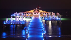 A waterfront restaurant with a long illuminated walkway leading to the dining area, surrounded by water. - Sueno Hotels Deluxe Belek - All Inclusive (Serik)