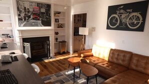 A living room with a brown leather sofa, a fireplace, a dining table with a laptop, and a painting of a motorcycle. - Duo Housing Hostel (Washington)