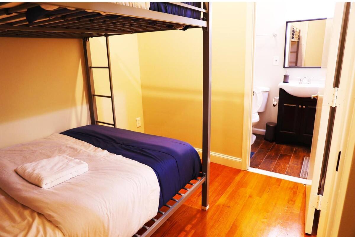 A bunk bed with a white and blue mattress, a wooden floor, and a bathroom with a sink and toilet visible through an open door.