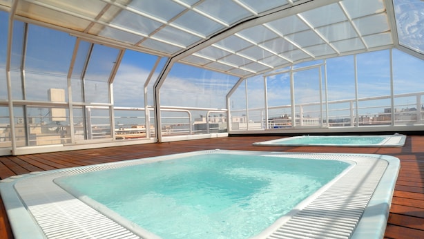 A glass-enclosed swimming pool area with clear blue water, wooden deck flooring, and a view of the cityscape through large windows. - Evenia President Hotel (Salou)