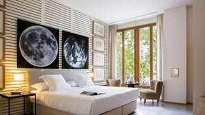Ein modernes Schlafzimmer mit einem großen Bett, einem Schreibtisch und einem Stuhl. An den Wänden hängen große Bilder und das Fenster bietet Blick auf Bäume.
