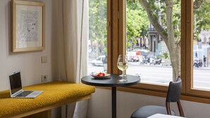 Ein Tisch mit einem Laptop, einem Weinglas und einem Teller Essen, platziert vor einem Fenster mit Blick auf eine Straße und Bäume.