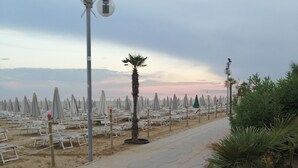 A beachside walkway with rows of beach chairs and umbrellas, a palm tree, and a street lamp. - Hotel Columbia & Ninfea (Jesolo)