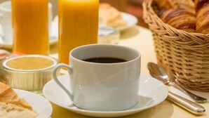 A breakfast setting with a cup of coffee, two glasses of orange juice, croissants, and a wicker basket. - Hôtel Le Home (Vincennes)