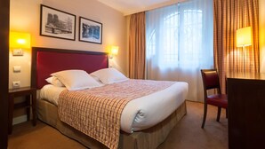 A hotel room with a large bed, two bedside lamps, a desk with a chair, and a window with curtains. - Hôtel Le Home (Vincennes)