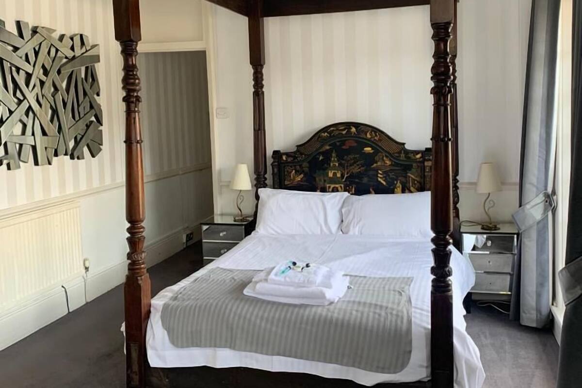 A four-poster bed with white linens and a folded towel, a nightstand with a lamp, and a decorative wall piece.