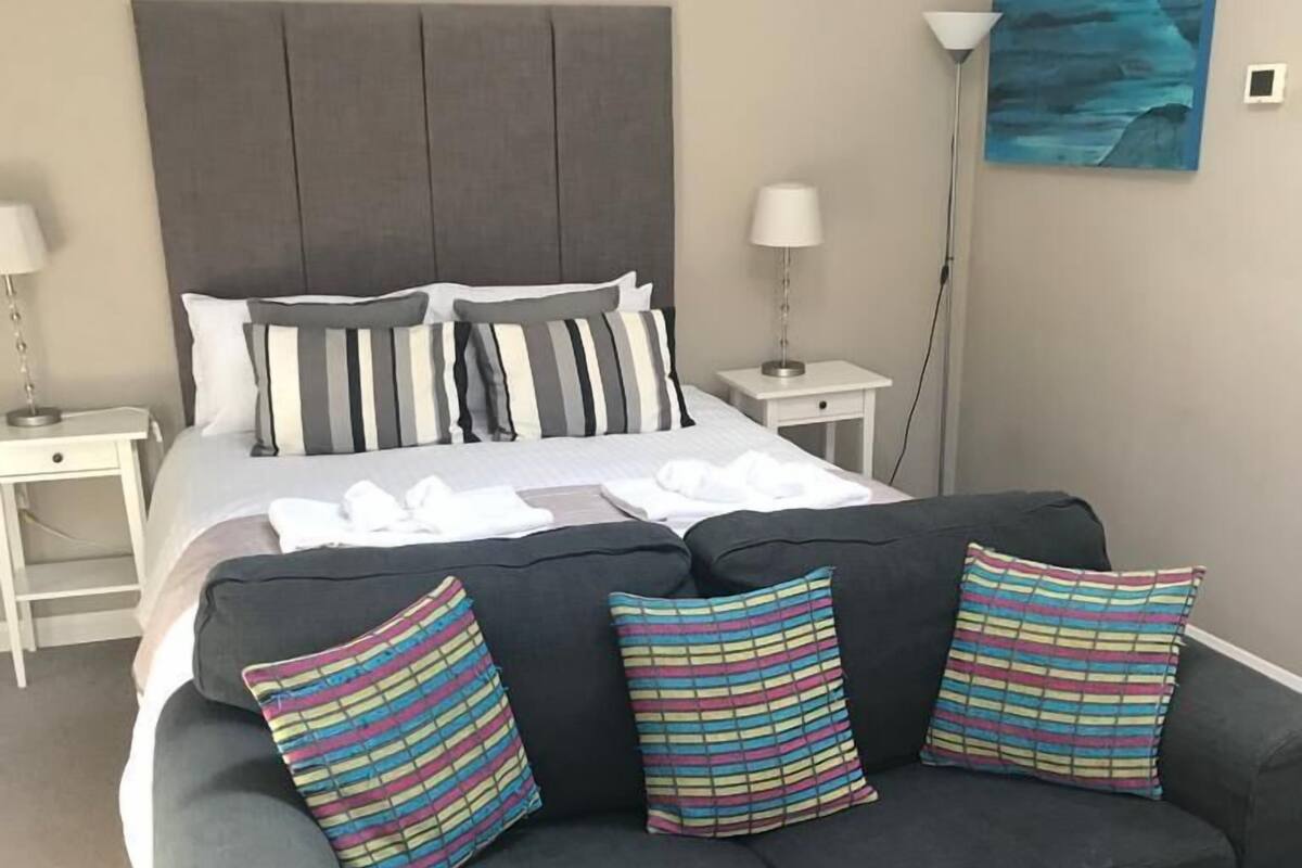 A modern hotel room with a large bed, a dark sofa with striped pillows, a bedside table with a lamp, and a wall painting.