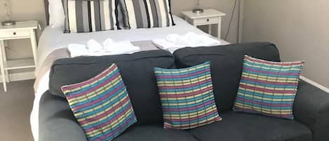A modern hotel room with a large bed, a dark sofa with striped pillows, a bedside table with a lamp, and a wall painting.