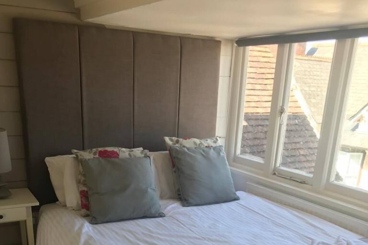A neatly made bed with pillows, a bedside table, and a window with a view of buildings.