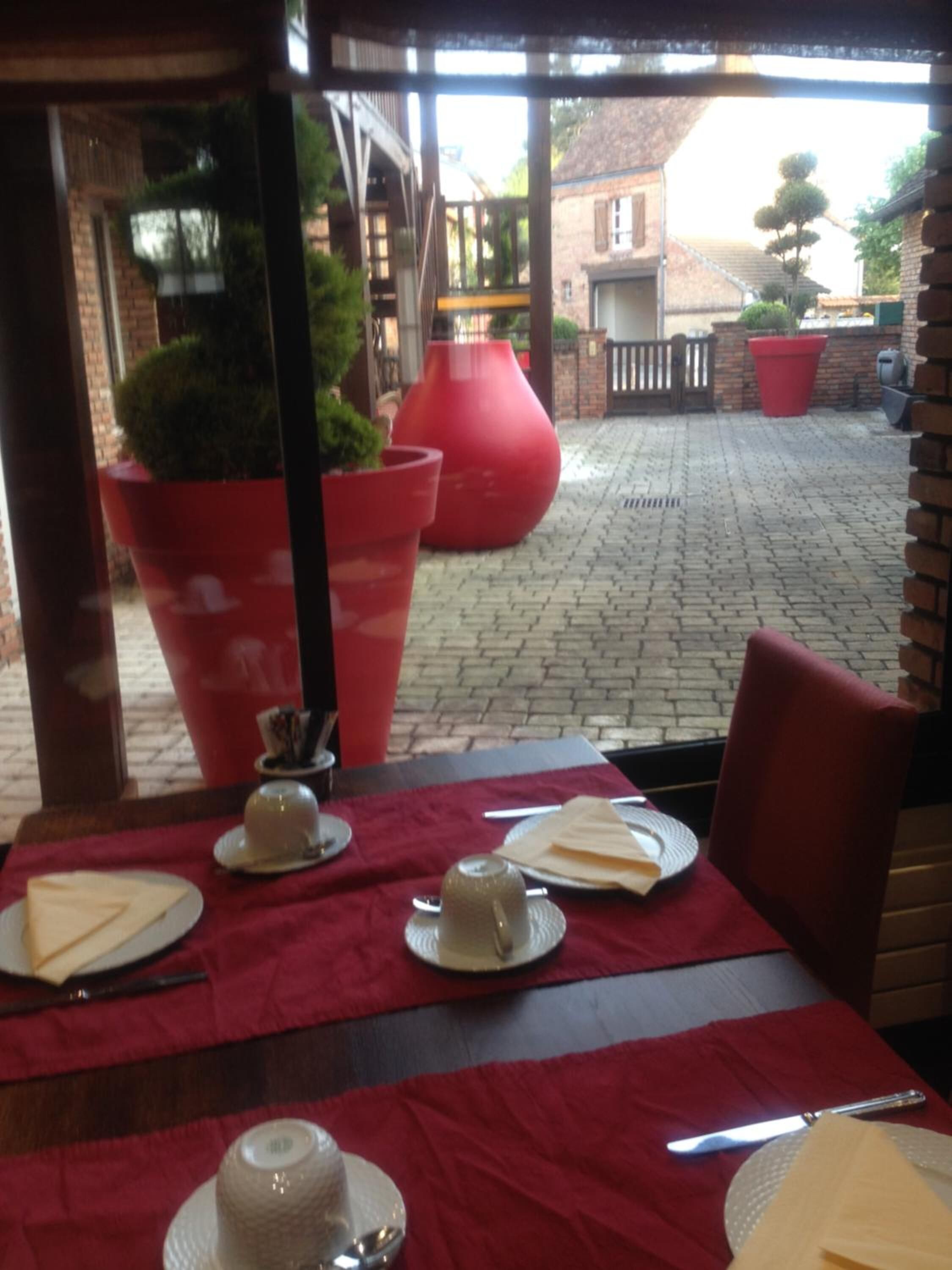Un área de comedor al aire libre con mesas preparadas para dos, plantas grandes en macetas y un camino de ladrillo que conduce a un edificio.